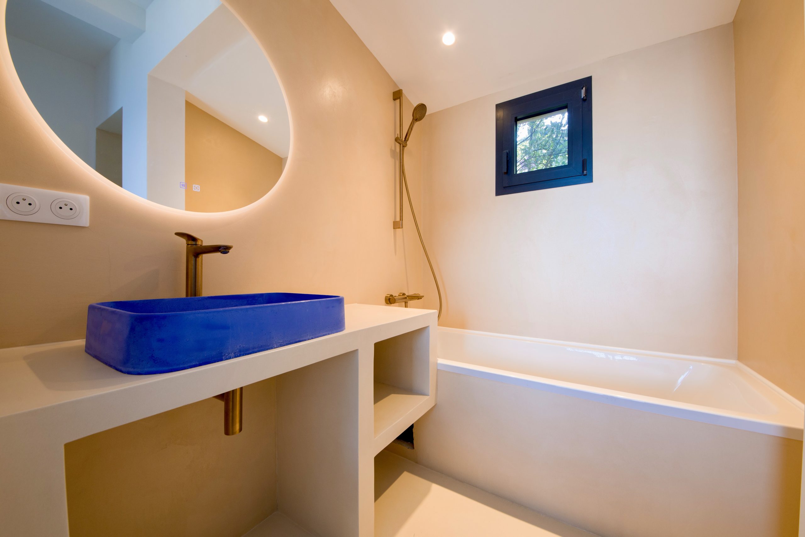 Salle de bain en béton ciré avec vasque bleue et miroir arrondi design à Hyères par Clavel Rénovation.